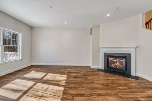 Bright living room with dark wood floors, large window bringing in sunlight, and a white-framed gas fireplace with mantel in-law suite addition Maine