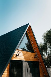 garage conversion to ADU Maine. The side of a triangular-roofed wooden house with a green metal roof, warm wood siding, and two lanterns, set against a clear blue sky.
